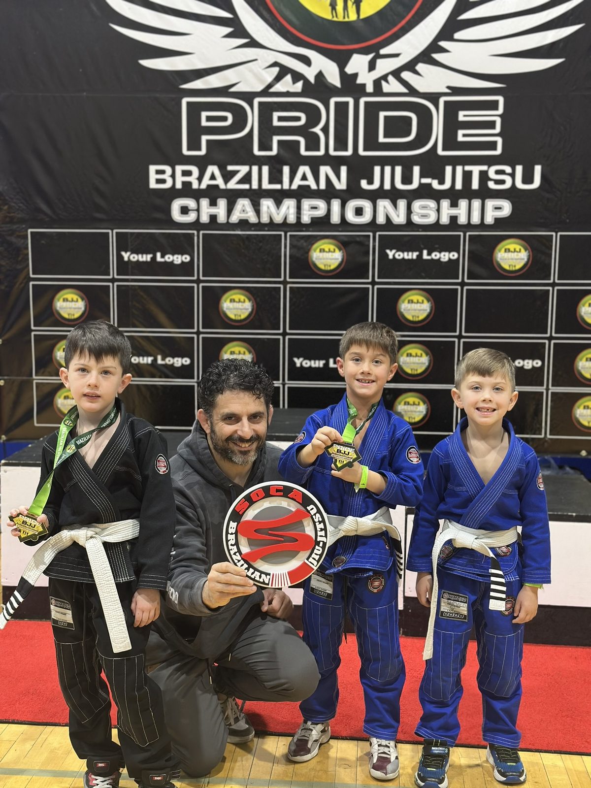 Young Soca BJJ competitors with their medals alongside their coach at a Brazilian Jiu-Jitsu championship
